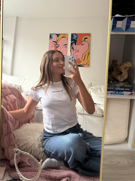 girl taking a mirror selfie wearing a white mirror moments t-shirt that says "you got this" and blue jeans in a bedroom with pink blanket